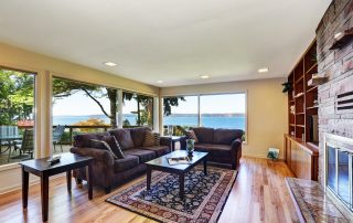 Interior of nicely furnished living room with large windows overlooking back deck. Has hardwood floor brown chocolate sofas persian style rug natural stone wall trim and a fireplace.
